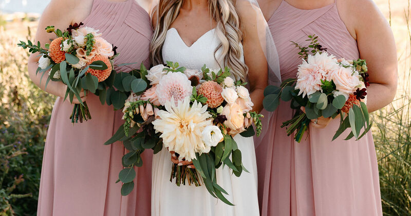 Bridal bouquet, bride’s bouquet with flowers done by Sweet Elegance Floral design florist in Kelowna, BC.
Hand tied bouquet of dahlias, roses, garden roses, spray roses and greenery in colours of pink, orange and yellow in a garden, rustic design.
