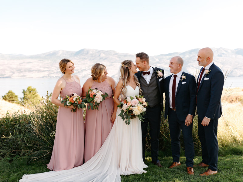 Wedding of Bride Kristina and Groom Paul, outdoor ceremony at Sanctuary Gardens in Kelowna, BC.  On a bright, clear day overlooking Lake Okanagan and vineyards.  Bride’s bouquet of pink, peach, orange and yellow roses and dahlias.  Rose petals down the aisle and ceremony centerpieces on wine barrels.  Bridesmaids in pink and groomsmen in suits.  Wedding photos taken by Tailored Fit Photography.  Wedding flowers done by Sweet Elegance Floral design, florist in Kelowna, BC.  