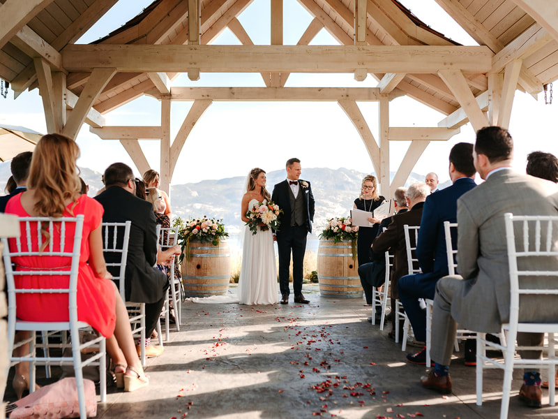Wedding of Bride Kristina and Groom Paul, outdoor ceremony at Sanctuary Gardens in Kelowna, BC.  On a bright, clear day overlooking Lake Okanagan and vineyards.  Bride’s bouquet of pink, peach, orange and yellow roses and dahlias.  Rose petals down the aisle and ceremony centerpieces on wine barrels.  Wedding photos taken by Tailored Fit Photography.  Wedding flowers done by Sweet Elegance Floral design, florist in Kelowna, BC.  